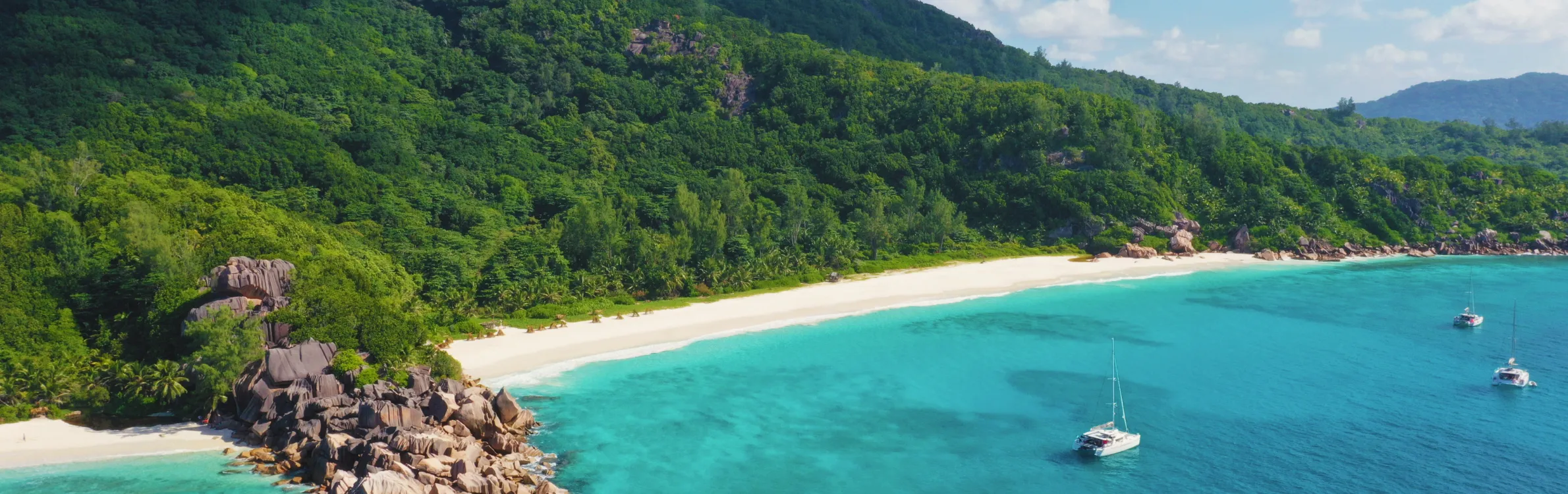 Langer Strand auf den Seychellen mit Booten im Meer und Wald im Hintergrund