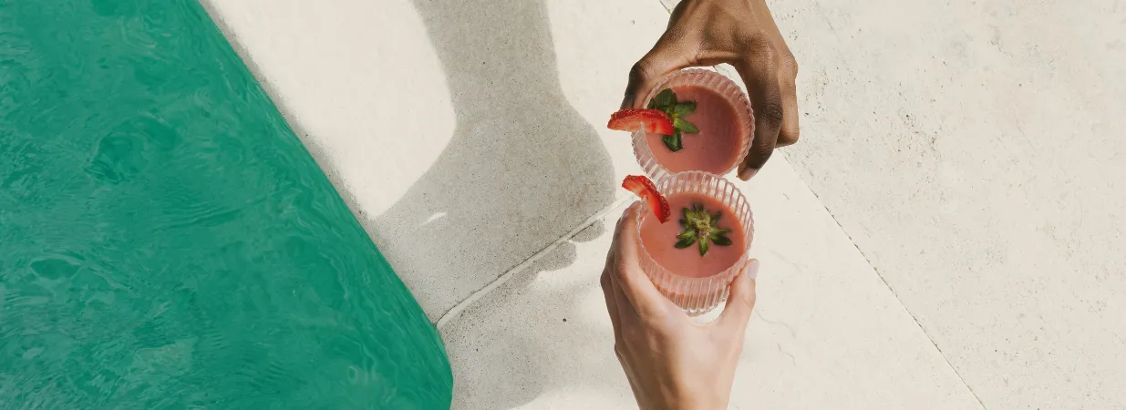 Zwei Hände halten Cocktails in der Hand neben dem Pool