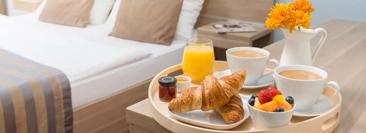 Frühstückskorb mit Croissant, Kaffee, Früchte neben dem Hotelbett